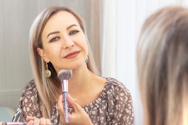 Une femme âgée met son maquillage. regarder dans le miroir. à moi-même un maquilleur. appliquer la poudre sur le visage avec un gros pinceau