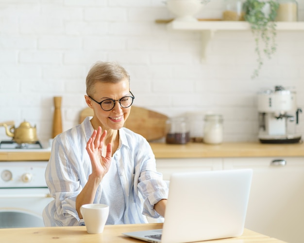 Une femme âgée moderne et heureuse portant des lunettes agitant une webcam sur un ordinateur portable tout en étudiant en ligne