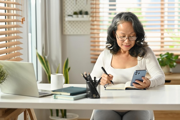 Femme âgée moderne vérifiant son plan de travail sur un téléphone intelligent et prenant des notes sur le bloc-notes