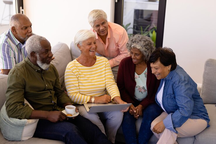 Photo femme âgée multiraciale tenant un ordinateur portable riant tout en étant assise avec des amis sur un canapé dans une maison de retraite. heureux, technologie sans fil, café, convivialité, soutien, vie assistée, concept de retraite.