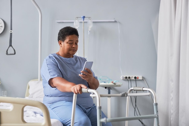 Femme âgée noire utilisant un téléphone portable assise seule dans une chambre d'hôpital
