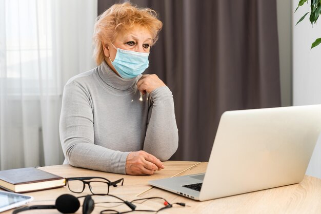 Une femme âgée portant un masque médical travaille sur un ordinateur portable à la maison. Communication avec les amis et la famille à distance pour les personnes âgées. Isolation. Quarantaine.