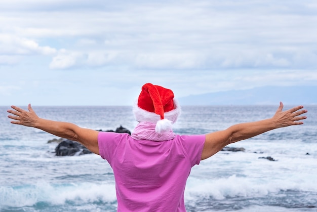 Femme âgée portant un masque de protection debout sur la plage à bras ouverts portant un bonnet de Noel