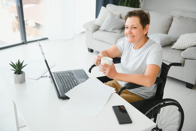 Femme âgée positive en fauteuil roulant travaillant sur ordinateur portable IT pigiste en ligne