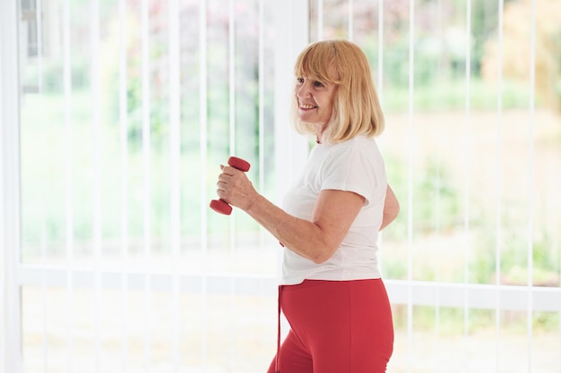 Femme âgée positive en vêtements sportifs à l'intérieur pendant la journée, faire des exercices avec des haltères