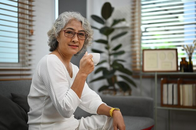 Femme âgée satisfaite femme assise sur un canapé confortable dans le salon avec une tasse de café Concept de style de vie de retraite