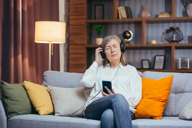 Femme âgée se relaxant à la maison le soir en écoutant un livre audio et un podcast en utilisant l'application sur son téléphone et ses écouteurs