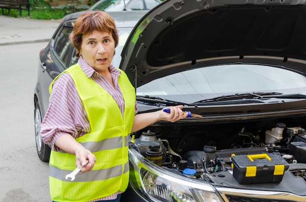 Une femme âgée se tient à côté d'une voiture endommagée avec un capot ouvert jeter leurs mains dans la confusion tenant des outils dans ses mains en attendant de l'aide