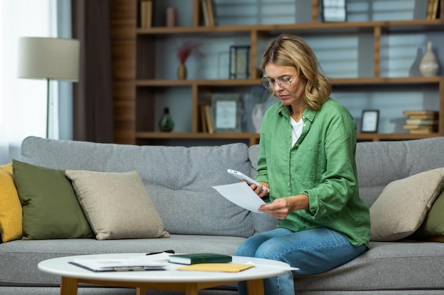 Femme âgée sérieuse et confuse dans des verres assis sur un canapé à la maison tenant avec inquiétude le téléphone et