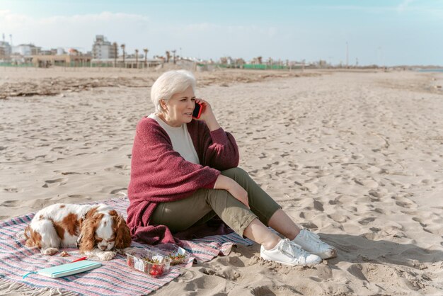 Femme âgée souriante assise sur la plage à côté de son chien endormi pendant la conversation téléphonique