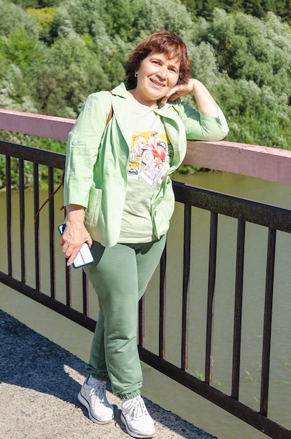 Une femme âgée souriante et attrayante se tient sur le pont par une journée ensoleillée d'été sur fond de nature