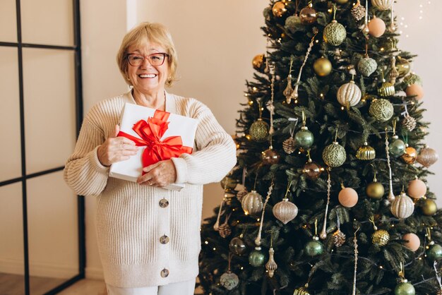 Une femme âgée souriante avec un cadeau de Noël près d'un arbre décoré