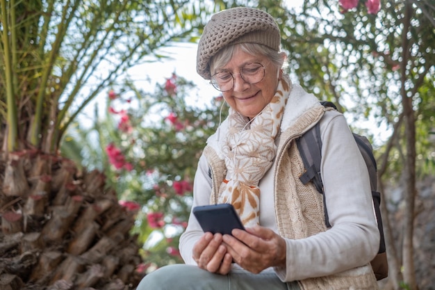 Femme âgée souriante avec casquette et sac à dos lors d'une excursion en plein air à l'aide d'un téléphone écoutant de la musique avec des écouteurs