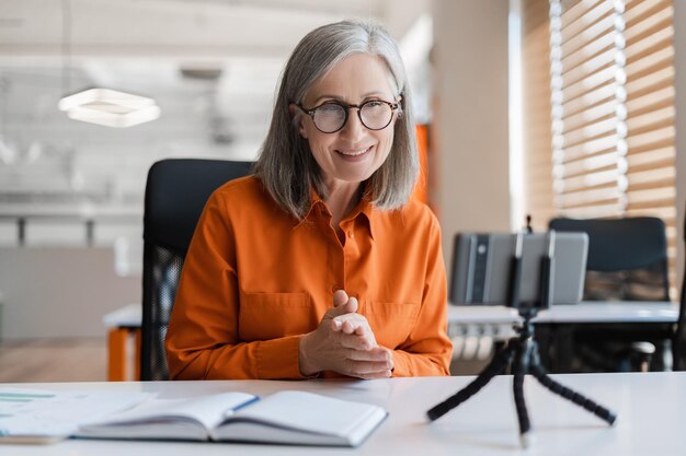 Femme âgée souriante et confiante utilisant la communication par téléphone portable en ligne ayant un appel vidéo Vidéo d'enregistrement réussie d'un influenceur aux cheveux gris regardant l'écran d'un smartphone assis sur le lieu de travail