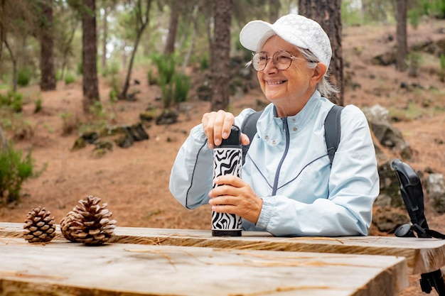 Femme âgée souriante lors d'une journée de randonnée dans les bois, assise à une table en bois pour se reposer et boire