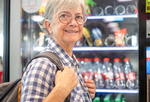 Femme âgée souriante avec des lunettes choisissant des collations et des boissons sur un distributeur automatisé gratuit 24h/24