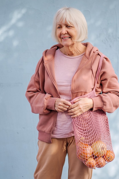 Femme âgée souriante en sweat à capuche confortable portant un sac en coton avec épicerie contre le mur bleu.