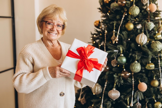Une femme âgée souriante tenant un cadeau près de l'arbre de Noël