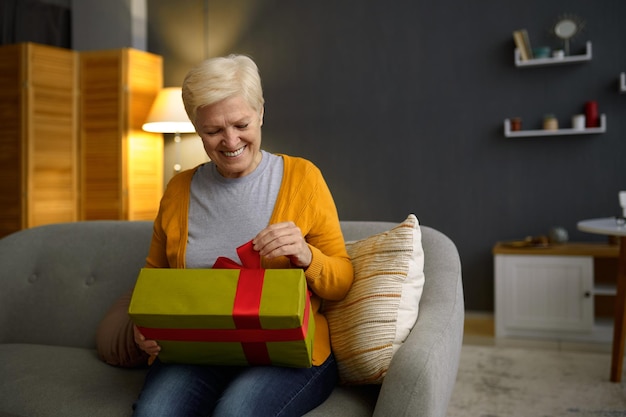 Une femme âgée tenant un paquet de boîtes-cadeaux avec un arc rouge assis à la maison salon