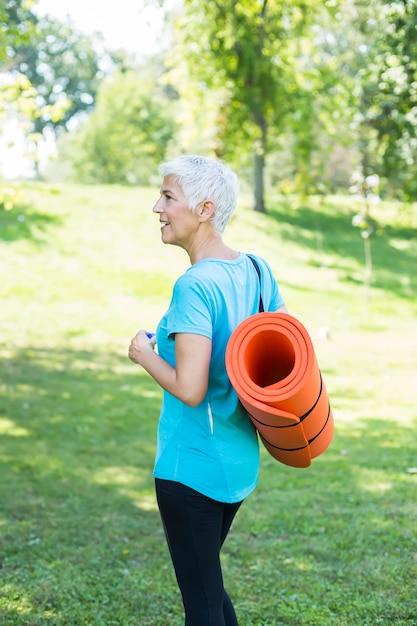 Femme âgée tenant un tapis de fitness sur le dos dans le parc et se préparant à faire de l&#39;exercice