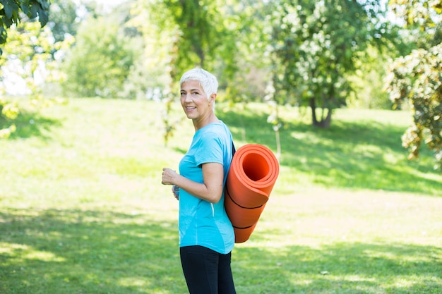 Femme âgée tenant un tapis de fitness sur le dos dans le parc et se préparant à faire de l&#39;exercice