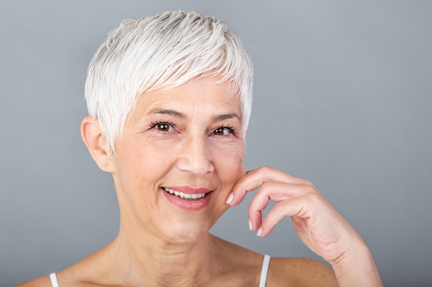 Une femme âgée tirant des joues pour ressentir la douceur et regardant la caméra Portrait de beauté d'une femme mature heureuse souriante avec les mains sur la joue isolée sur un fond gris Processus de vieillissement et concept de peau