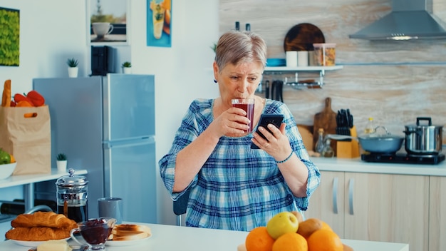 Femme âgée utilisant un gadget mobile dans la cuisine. Personne âgée authentique utilisant la technologie Internet moderne pour smartphone. Communication en ligne connectée au monde, temps libre avec gadget à r