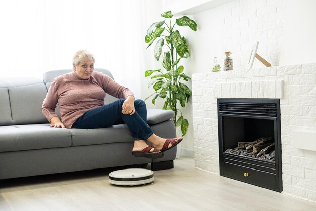 Une femme âgée utilise un robot aspirateur dans un appartement.