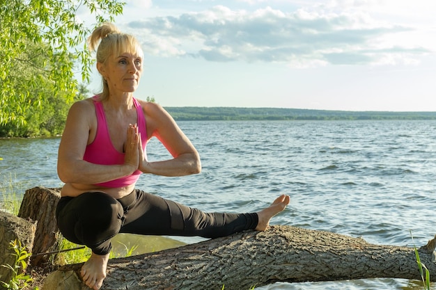 Une femme âgée en vêtements de sport pratiquant le yoga debout dans un demi-pas et tenant ses mains jointes namaste sur un arbre au bord du lac Sports de plein air Yoga dans la vieillesse