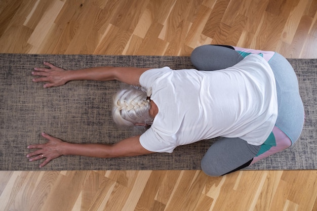 Femme agenouillée sur un tapis et faisant une asana balasana tout en pratiquant le yoga à la maison