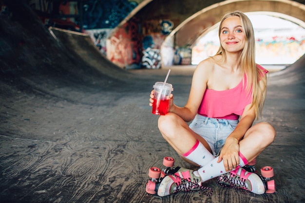 Femme agréable et positive est assise avec ses jambes croisées et tenant une tasse en plastique avec du jus rouge