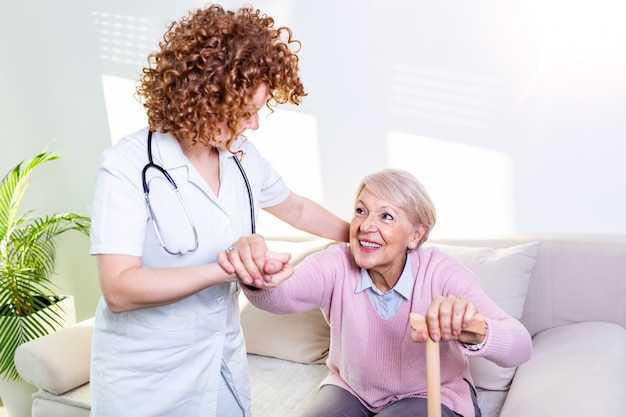 Femme Aidant Aidant Une Femme âgée à Se Lever Du Canapé Dans Le Salon. Infirmière Souriante Aidant Une Femme âgée à Se Lever.