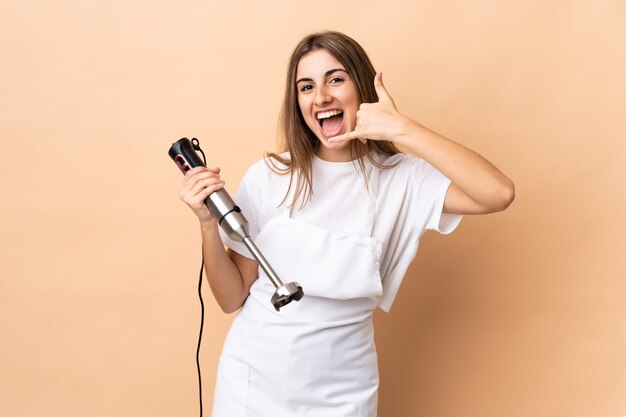 Femme à l'aide d'un mixeur à main sur un mur isolé faisant un geste de téléphone. Rappelez-moi signe