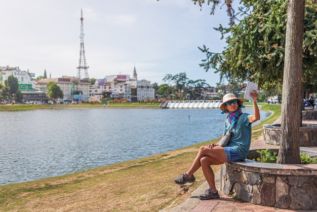 Femme à l'aide de téléphone intelligent tablette dans le parc de la ville verte du lac à la ville de Da Lat, Vietnam. Dame avec chapeau vietnamien prenant selfie