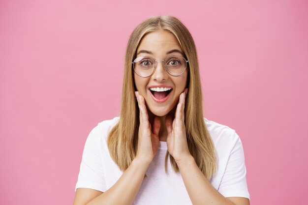 Femme aimant de nouvelles lunettes choisies en magasin se sentir heureux de trouver le cadre droit touchant les joues du bonheur souriant joyeusement à la caméra portant des lunettes transparentes et un t-shirt sur un mur rose