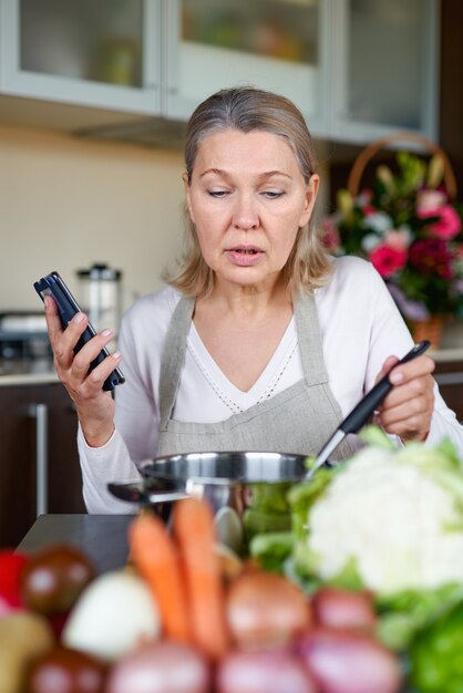 Femme aînée, dans, cuisine, préparer, nourriture, et, tenue, smart phone