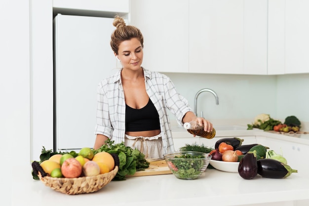 Femme ajoutant de l'huile d'olive extra vierge à sa salade pendant la cuisson dans une cuisine blanche