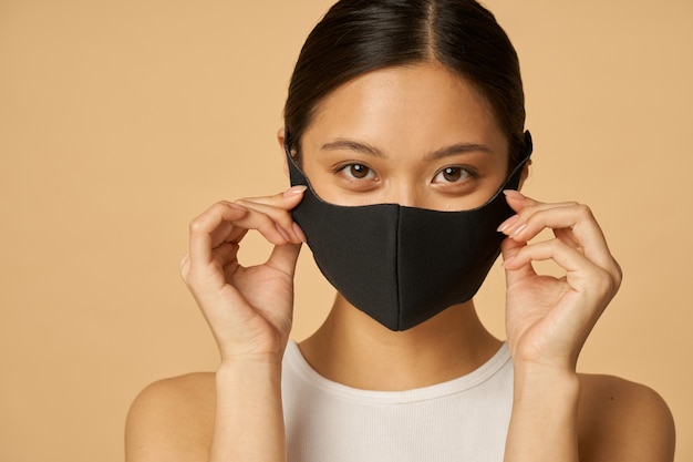 femme ajustant le masque facial noir et regardant la caméra, posant isolée sur fond beige.