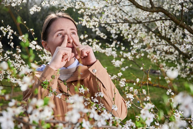Femme allergique souffrant d'une allergie saisonnière au printemps posant dans un jardin en fleurs au printemps Jeune femme éternuant devant un arbre en fleurs Concept d'allergie de printemps