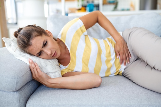 Photo femme allongée sur un canapé à la recherche de malades dans le salon belle jeune femme allongée sur le lit et se tenant la main sur le ventre