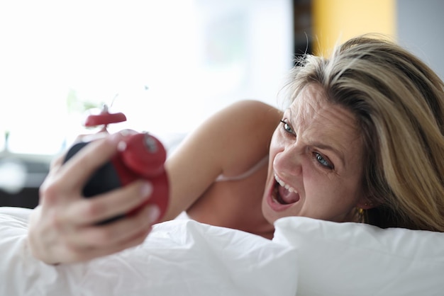 Photo femme allongée dans son lit avec un réveil dans les mains et criant