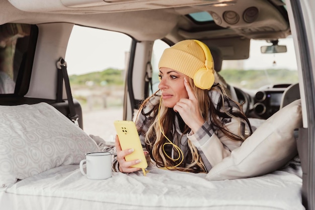 Femme allongée face contre terre sur le lit de son camping-car tout en utilisant un téléphone portable pour écouter de la musique avec des écouteurs