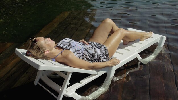 Femme allongée sur un lit de bronzage avec des lunettes de soleil et un châle en soie bohème Une fille se repose sur une jetée sous-marine en bois d'inondation Le trottoir est recouvert d'eau dans le lac