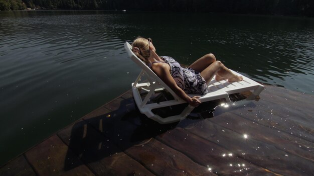 Femme allongée sur un lit de bronzage avec des lunettes de soleil et un châle en soie bohème Une fille se repose sur une jetée sous-marine en bois d'inondation Le trottoir est recouvert d'eau dans le lac