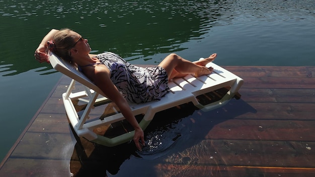 Femme allongée sur un lit de bronzage avec des lunettes de soleil et un châle en soie bohème Une fille se repose sur une jetée sous-marine en bois d'inondation Le trottoir est recouvert d'eau dans le lac