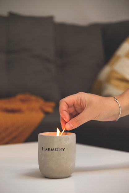 Une femme allume une bougie dans un chandelier en béton sur une table dans le salon à côté d'un canapé avec des oreillers et un bl...