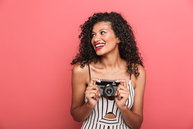 femme américaine joyeuse aux cheveux bouclés photographiant sur un appareil photo rétro isolé sur un mur rose