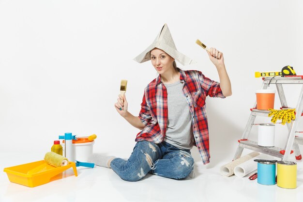 Femme amusante au chapeau de journal assis sur le sol avec une brosse, instruments pour la rénovation de la chambre d'appartement isolée sur fond blanc. Papier peint, accessoires pour le collage, outils de peinture. Réparer le concept de maison.