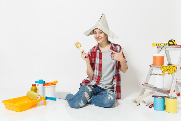 Femme amusante au chapeau de journal assis sur le sol avec une brosse, instruments pour la rénovation de la chambre d'appartement isolée sur fond blanc. Papier peint, accessoires pour le collage, outils de peinture. Réparer le concept de maison.