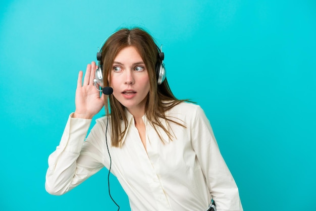 Femme anglaise de télévendeur travaillant avec un casque isolé sur fond bleu écoutant quelque chose en mettant la main sur l'oreille